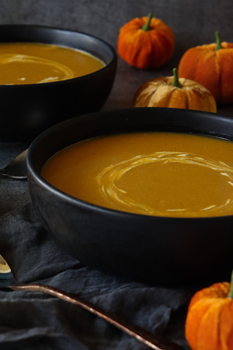 Stock photo showing close-up view of homemade Halloween pumpkin soup in bowls on dark grey muslin next to a metal spoon and plush fabric pumpkins. The soup has been garnished with swirls of yoghurt.