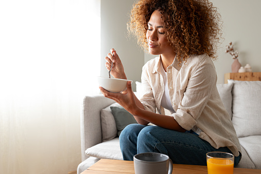 Young relaxed pensive African American woman eating healthy breakfast sitting on the couch at home cozy living room. Copy space. Lifestyle concept.
