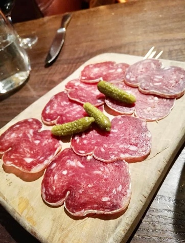 This enticing charcuterie board is a masterpiece of flavors and textures. A carefully arranged selection of salami, saucisson sec, and other savory delights awaits on a rustic wooden serving board. The artful presentation, accompanied by gherkins and pickles, enhances the visual appeal of this culinary creation. With a knife poised for slicing and a glass in the background, it invites a leisurely and indulgent dining experience at a charming restaurant.