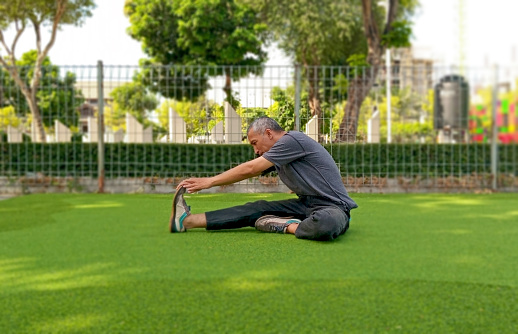 Asian man in positive facial expression is stretching in the park.