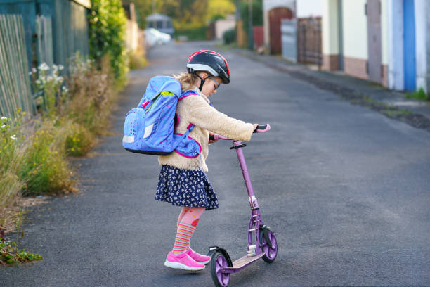 little girl rides a kick scooter on her way back to school. cute child with backpack and with safety helmet. happy schoolchild. - helmet bicycle little girls child imagens e fotografias de stock