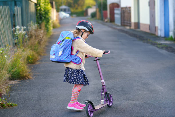 Cтоковое фото Маленькая девочка едет на самокате по дороге в школу. Симпатичный ребенок с рюкзаком и в защитной каске. Счастливый школьник.