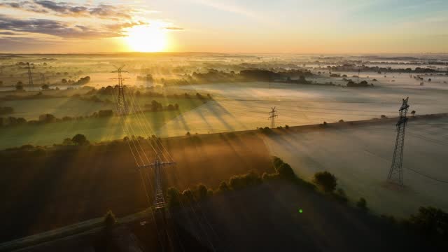 Aerial footage over industry utility power lines and pylon structure in foggy agricultural hedge landscape by the morning sunrise. Multiple power lines and pylons stretch across the farmland.