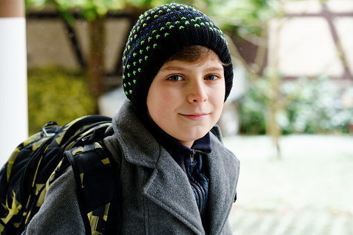 Portrait of preteen kid boy outdoors. Handsome child boy in coat and hat in winter. Schoolchild on the way to school with backpack satchel during snow.