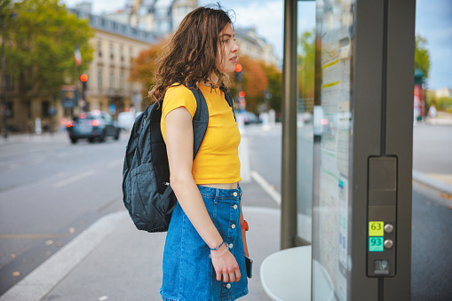 Young woman traveling solo, standing on bus stop in the city and waiting for public transportation