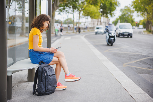 Young woman on solo adventure, sitting on bus stop and waiting for public transportation, checking the sights on her smart phone