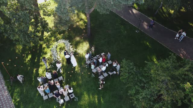 Aerial top view of wedding ceremony in the park with arch decorated with white flowers and the the guests stand and applaud the bride as she goes to the groom , unrecognisable people.