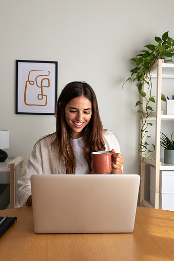 Happy young woman working at home office having coffee. Caucasian female entrepreneur using laptop. Vertical image. Working at home concept.