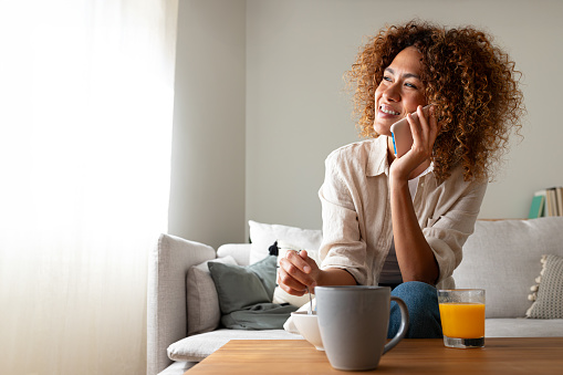Young multiracial hispanic woman talking on the phone while having breakfast sitting on the sofa at home cozy living room. Copy space. Coffee and technology concept.