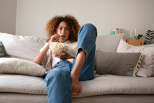 Young multiracial woman watching movie at home eating popcorn, sitting on the couch with surprise expression. Leisure activity.