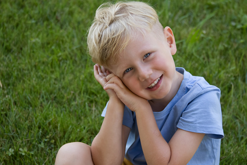 A blond boy in a blue T-shirt  laughs against the backdrop of a green lawn copy space