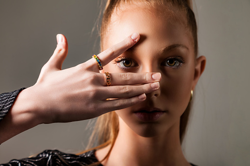 Perfect chic teen girl model looking eye through fingers at camera, studio shot. Portrait of lovely teenage cover lady 13 year old posing with gesture. Fashion image style concept. Copy ad text space