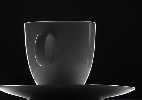 Porcelain coffee cup on a saucer isolated on black background. Close up. Dark atmosphere. Backlit studio scene. Ceramic.