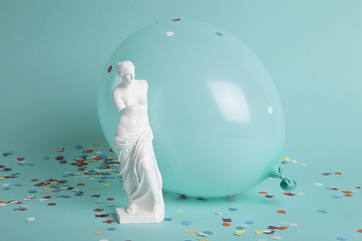 a venus in front of a balloon of the same color as the background. Confetti on the ground. color gradation. Minimalist, trendy still life photography.