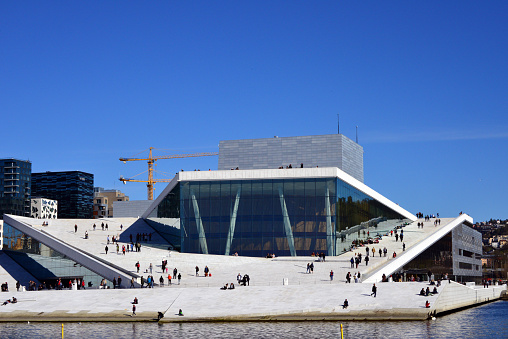 Oslo, Norway: Oslo opera house reflected on the Bjørvika Bay waterfront with people strolling on the open roof plaza - The building, a two-part theater with opera and ballet, was modeled on a floating iceberg, was designed and built by the Norwegian architectural firm Snøhetta (“Snow Cap”), which had already designed the new library of Alexandria in Egypt - with Christoph Kapeller as design leader and project director - Bjørvika neighborhood in the Sentrum borough, named after Bjørvika bay.