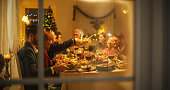 Happy Family Gather at Home for a Traditional Christmas Dinner with a Turkey Roast. Living Room is Crowded with Senior and Young Adults Enjoying Holiday Dishes. Creative Shot Through a Snowy Window