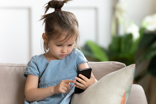 Smart little girl sit on couch at home using modern smartphone gadget, concentrated serious cute small preschooler daughter kid addicted to new technology, surfing on cellphone device