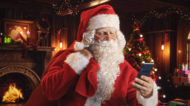 istock Happy Santa Claus with White Fluffy Beard and a Red Costume is Using a Smartphone. Accepting Online Gift Orders From All the Good Kids Around the World. Old Santa Holding a Bag with Presents 1744506684