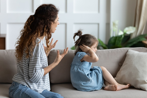 Angry young mother sit on couch talking lecturing offended little preschooler daughter, small girl child close cover ears ignore mad mom scolding, avoid family conflict or misunderstanding