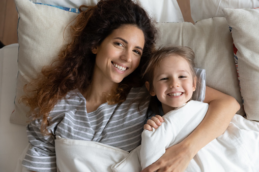 Top view portrait of smiling young mother and cute little daughter lying in bed under blanket ready to sleep, happy millennial mom or nanny hug cuddle in bedroom with small preschooler girl child