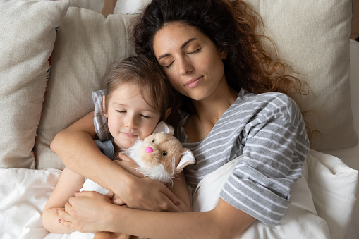 Top view of calm young mother lying relaxing in cozy bed with cute little daughter, millennial mom hug cuddle with small preschooler girl child sleeping together in comfortable home bedroom