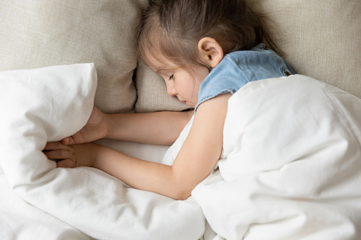 Top view of little preschooler girl lying sleeping peacefully on soft fluffy pillow covered with warm duvet, calm small child fall asleep take nap on comfortable mattress in cozy bed in home bedroom