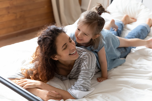 Overjoyed young mom lying on bed playing with cute small preschooler daughter together, happy millennial mom or nanny relax have fun in bedroom with little girl child, enjoy weekend at home