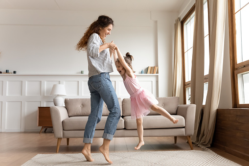 Happy young mother dancing swirling with excited little preschooler daughter relaxing together in living room, overjoyed millennial mom or nanny have fun with small girl child playing at home