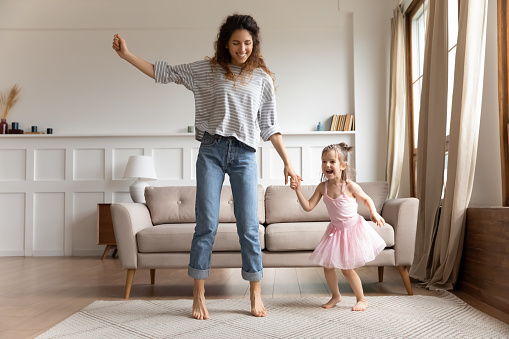 Overjoyed millennial mother and small preschooler daughter dance jump together in cozy living room at home, happy young mom or nanny have fun with cute little girl child, relax listen to music