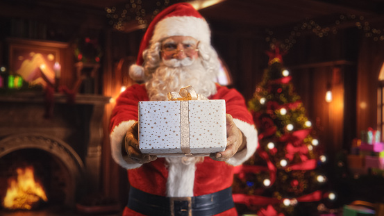 Portrait of a Happy Santa Claus Offering Beautifully Wrapped White Gift To Children. Old Man Looking at Camera, Smiling While Giving the Present Away. Concept of Christmas and New Year's Celebrations