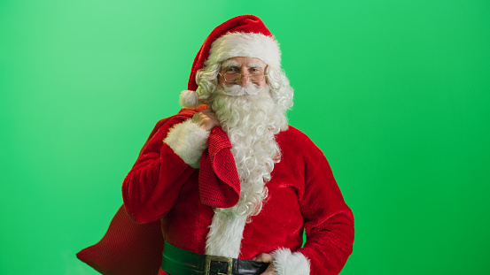 Studio Shot Isolated Mockup Template: Jolly Santa Claus Standing On Green Screen Chromakey Background And Holding Bag With Gifts, Looking at Camera. Christmas, New Year, Holiday Celebration