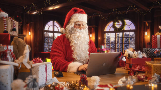 istock Arc Shot of Father Christmas Sitting and Using a Modern Laptop Computer. Santa is Browsing Online, Communicating with Clients, Managing a Successful Online Shopping Business with Gifts for Children 1744549652