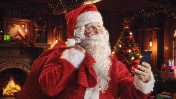 istock Happy Santa Claus with White Fluffy Beard and a Red Costume is Using a Smartphone. Accepting Online Gift Orders From All the Good Kids Around the World. Old Santa Holding a Bag with Presents 1744549669