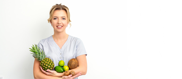 Portrait of a young caucasian smiling female nutritionist with different fruits in her hands over white background