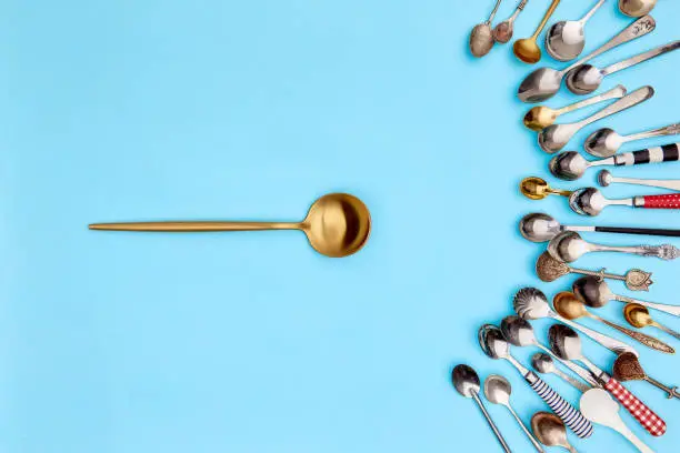 Tips of cutlery. Top view photo one soup-spoon surrounded of variety of antique silverware and gold spoons against blue studio background. Concept of food, holiday, table setting, retro, vintage