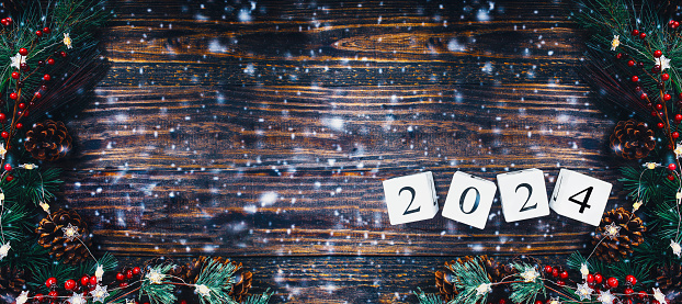 New Year's 2024 wood calendar blocks banner. Christmas tree lights, pine branches, red winter berries and snow over wooden table background. Top view, flat lay with copy space available.