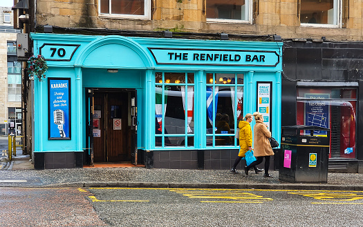 Exterior view of the brightly blue-colored Renfield Bar, sited at 70 Renfield St, Glasgow G2 2RA.  The flags of the Union, England, Scotland, Wales and Northern Ireland are visible in the Window.  2  people are walking past.