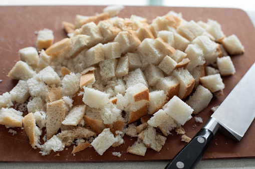 Closeup view of a brown cutting board with white bread cut in small square pieces and a kitchen knife on the side