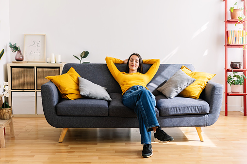 Full angle view of young pretty woman relaxing on cozy sofa with hands over head at modern bright apartment. Calmness and wellfare concept.