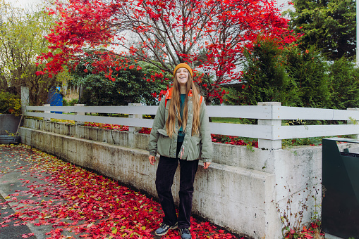 Front view of a happy female in red jacket with backpack walking in the city contemplating the bright red trees with maple leaves falling on the ground during autumn season in Norway