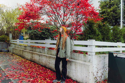 Side view of a happy female in red jacket with backpack walking in the city contemplating the bright red trees with maple leaves falling on the ground during autumn season in Norway