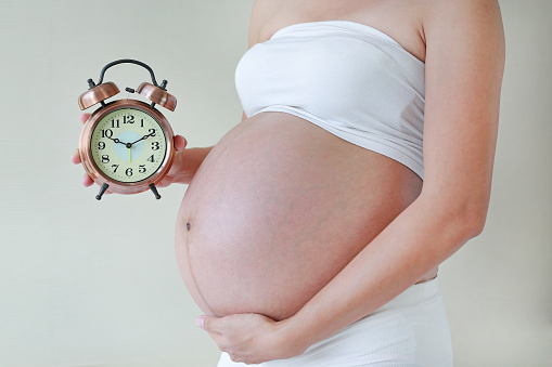 Pregnant woman with big belly in white underwear holding vintage alarm clock. Pregnancy waiting baby concept.