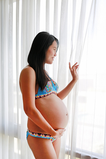 Pregnant Asian woman standing near window at home. Woman in underwear.