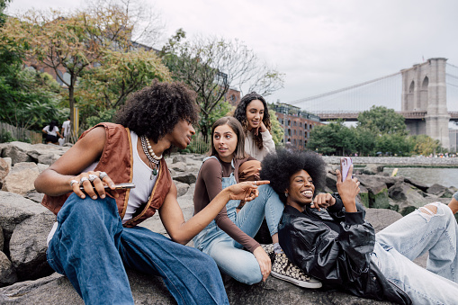 Multi-ethnic group of friends resting and enjoying a break next to Brooklyn Bridge in New York. They are enjoying the awesome view and texting on mobile.