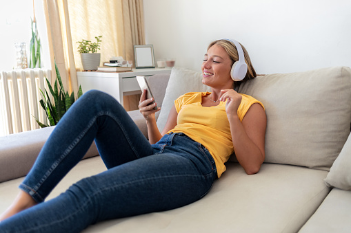 Shot of a young woman using a smartphone and headphones on the sofa at home.