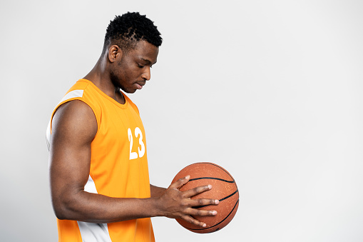 Serious muscular African man wearing orange sportswear playing basketball, holding ball isolated on white background. Sport competition concept