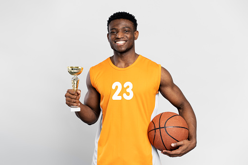 Confident smiling African man, basketball player holding trophy cup and ball, celebration success looking at camera isolated on white background. Sport victory, winning competition, awarding concept