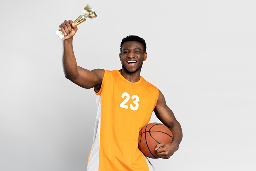 Portrait of overjoyed African man, basketball player holding ball and trophy cup, celebration success isolated on white background. Sport victory, winning competition concept