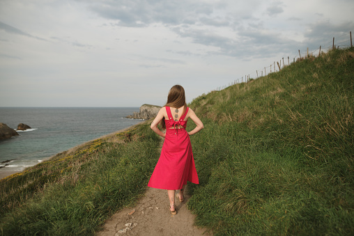 Rear view of a young woman with a red dress walking a path by the sea