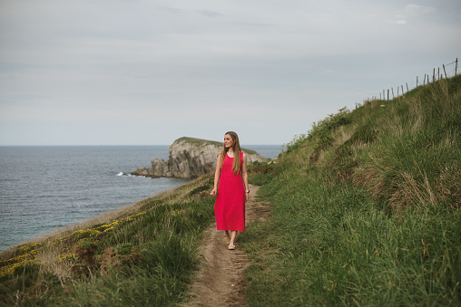 A young woman with red dress walking a path by the sea